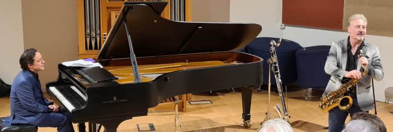 Jason Rebello seated at the piano, with Tim Garland holding his saxophone and speaking to the audience.