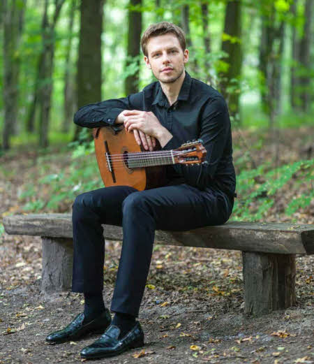 Michael is seated on a wooden bench in light woodland, his arms resting on his guitar which is on his lap.