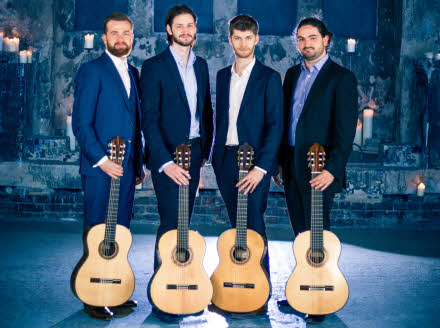 The four guitarists standing in a row with their guitars held down in front of them.