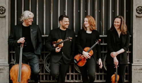 The Dante Quartet standing in a line with their instruments in front of an iron gateway. They are wearing black and looking cheerful.