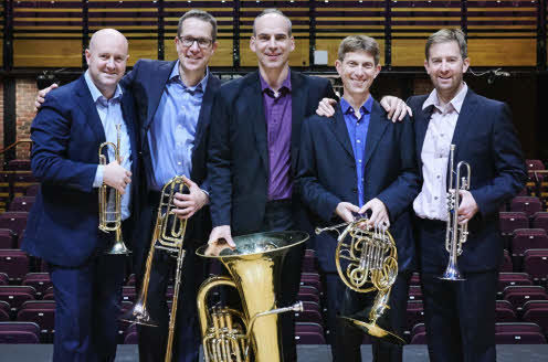 The five members of the quintet, holding their instruments and standing in a row on a stage with their backs to the concert hall seating