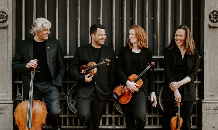 The Dante Quartet standing in a line with their instruments in front of an iron gateway. They are wearing black and looking cheerful.