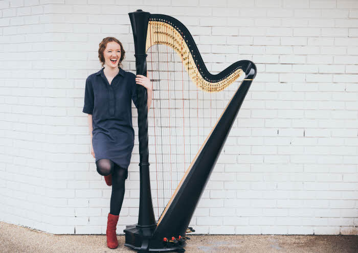 Alis is standing with her left arm wrapped around her harp whilst her right leg is bent at the knee and leaning against a white-washed brick wall