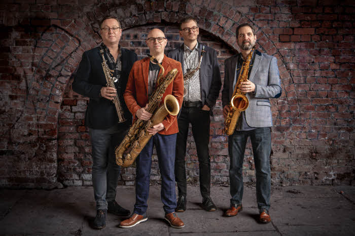 The quartet standing with their instruments on a pavement in front of the bricked-in arches of a bridge