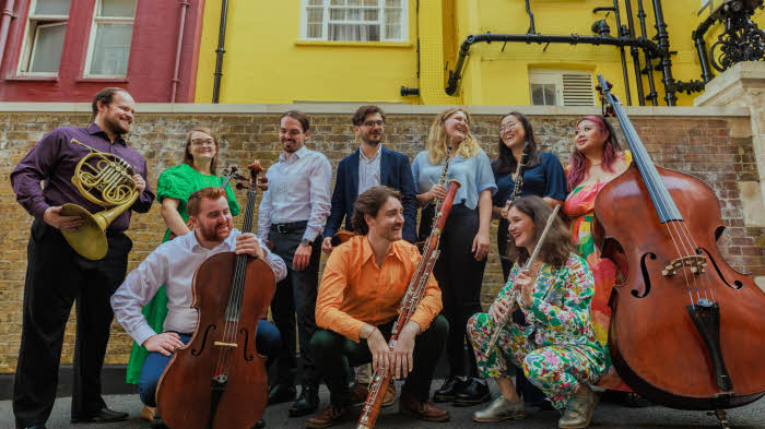 The Collective standing or crouching with their instruments in two rows in front of a brick wall, behind which are the brightly painted back walls of two terraced houses.