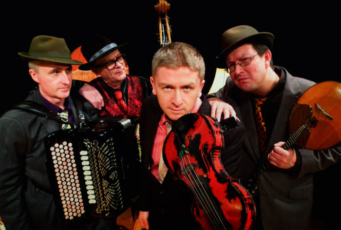 The four players in a tight group, holding their instruments, lean forward towards the camera with a mock-scowl.